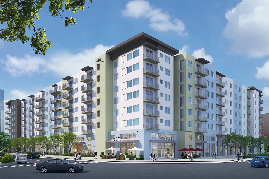 Multi-story residential apartment building exterior with white and sage green facade, dark roof overhangs, retail storefronts at street level, parked cars, blue sky with clouds.