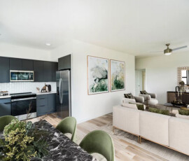 Open-concept apartment interior with dark kitchen island, white subway tile backsplash, beige living room seating, green accent chairs, artwork, wood-style flooring throughout.