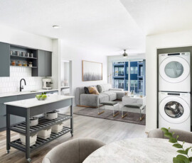 Apartment living space with dark charcoal kitchen on left, white appliances, open shelving, gray sofa in living area, stacked washer-dryer in alcove, gray accent rug, sliding doors with blue exterior.