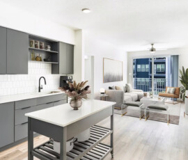 Contemporary apartment with dark gray cabinetry, white countertops, open shelving with decorative items, white subway backsplash, gray upholstered seating in living room, large windows with blue exterior views.